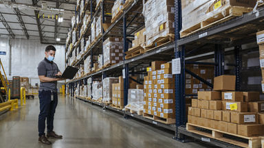 Male wearing a mask and using a laptop in a warehouse full of boxes