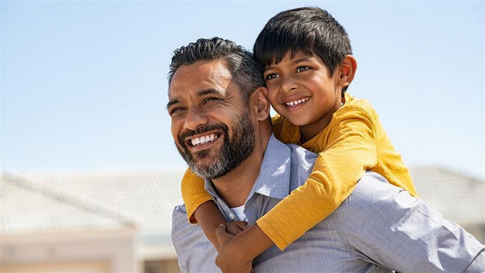 Happy middle eastern child enjoying ride on father back outdoor. Smiling dad giving piggyback ride to son on street while looking away with copy space. Indian cheerful man carrying on shoulder kid.