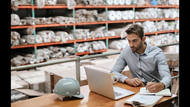 Manager checking inventory and online orders on a laptop while sitting at a desk in a carpet warehouse with shelves of stock in the background; Shutterstock ID 1414684886; purchase_order: New Web Images; job: ; client: ; other: 