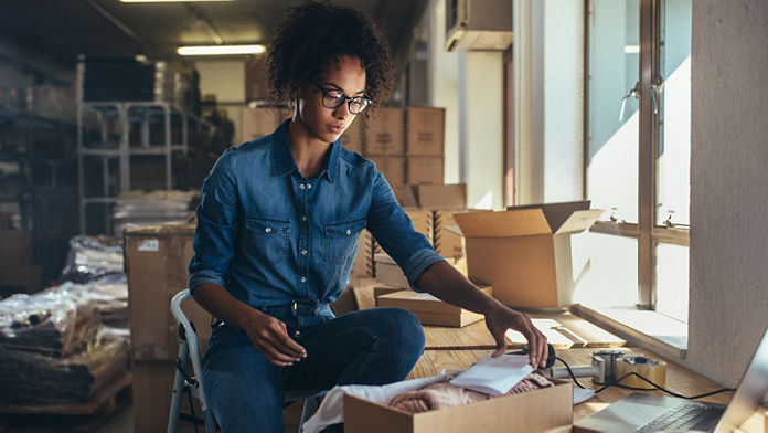 Woman picking up piece of paper from a box with a laptop in front of her in store room setting